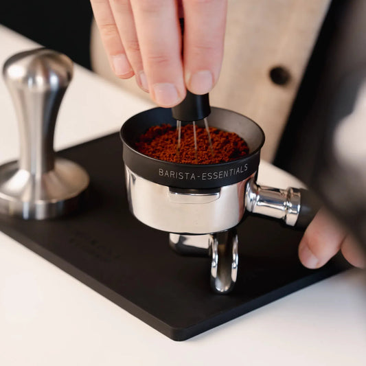 Hand verdeelt koffie met een WDT-tool boven een zwarte mat