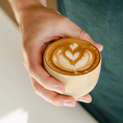 Zandgeel cappuccino kopje van keramiek met latte art, vastgehouden door barista. Ideaal voor thuisgebruik met populaire espressomachines en melkopschuimers.