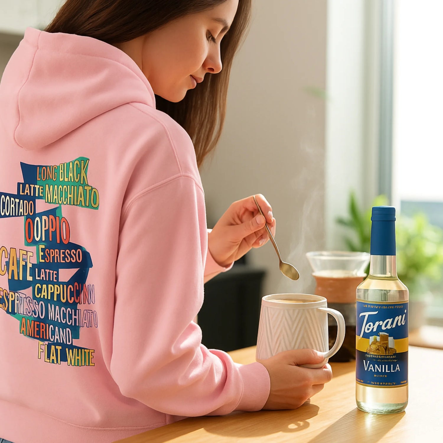 Vrouw in roze koffiehoodie maakt koffie met Torani suikervrije vanillesiroop – perfect als cadeau voor de bewuste koffieliefhebber