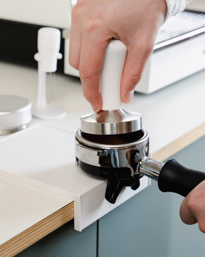 Een portafilter en tamper boven de kleine witte tampermat met hoek