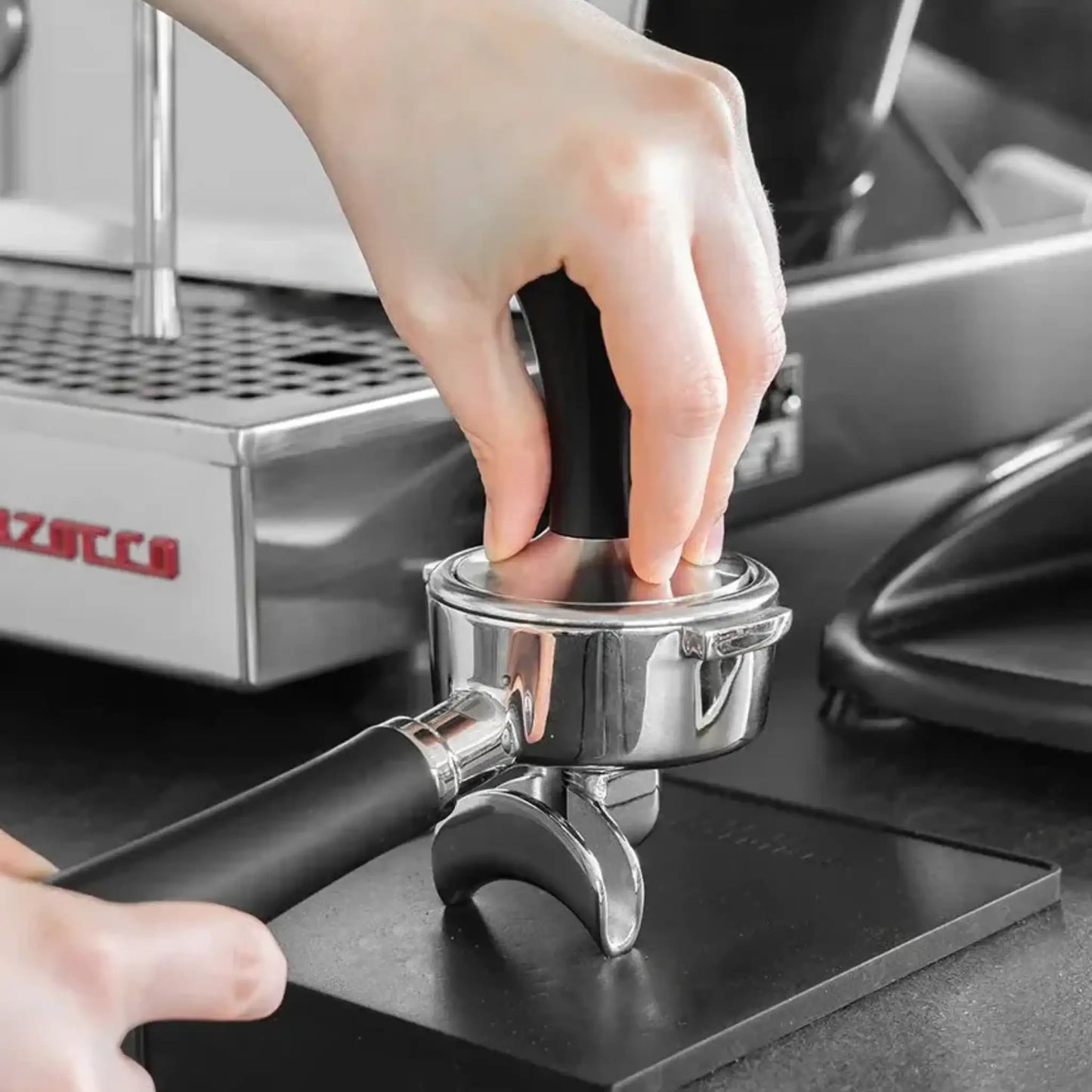 Thuisbarista drukt gemalen koffie aan met een zwarte espresso tamper in een portafilter.