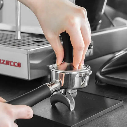Thuisbarista drukt gemalen koffie aan met een zwarte espresso tamper in een portafilter.