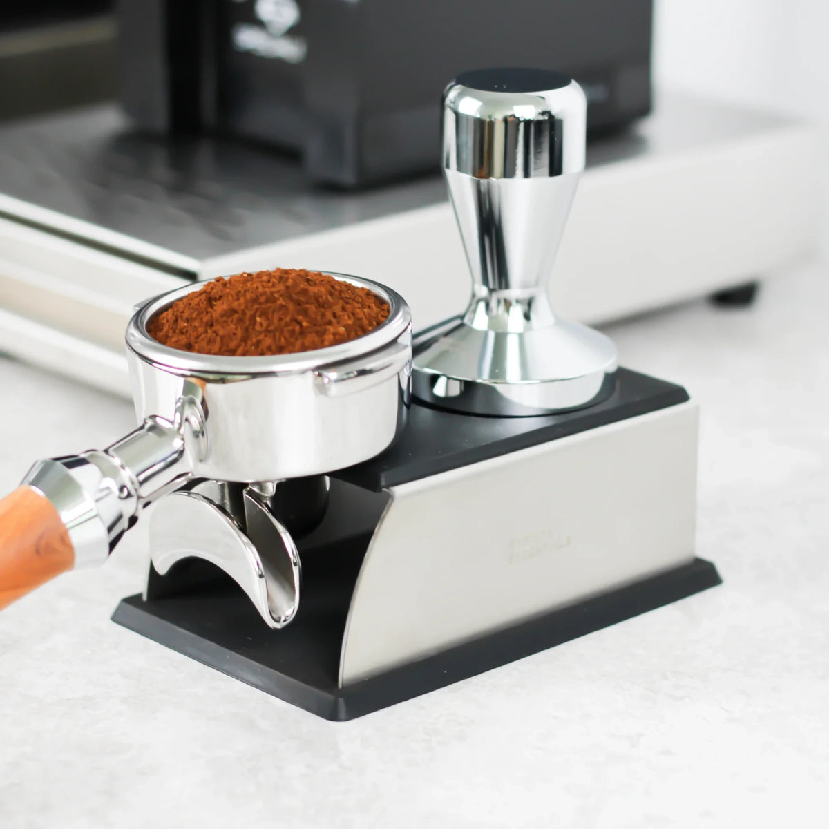 Gevulde portafilter en tamper op een zilveren tamping station naast een koffiemolen