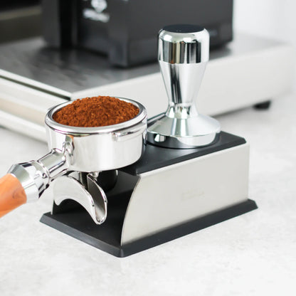 Gevulde portafilter en tamper op een zilveren tamping station naast een koffiemolen