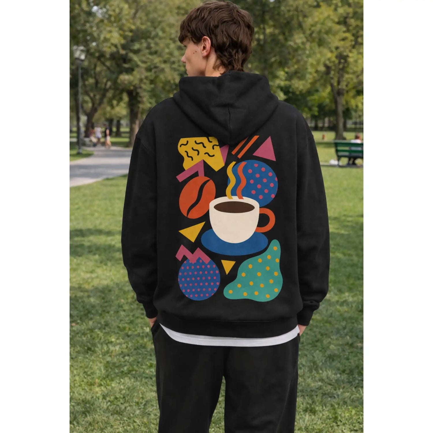 Zwarte koffie hoodie met abstracte koffie illustratie en kopje koffie op de rug, gedragen door een koffiefan in het park