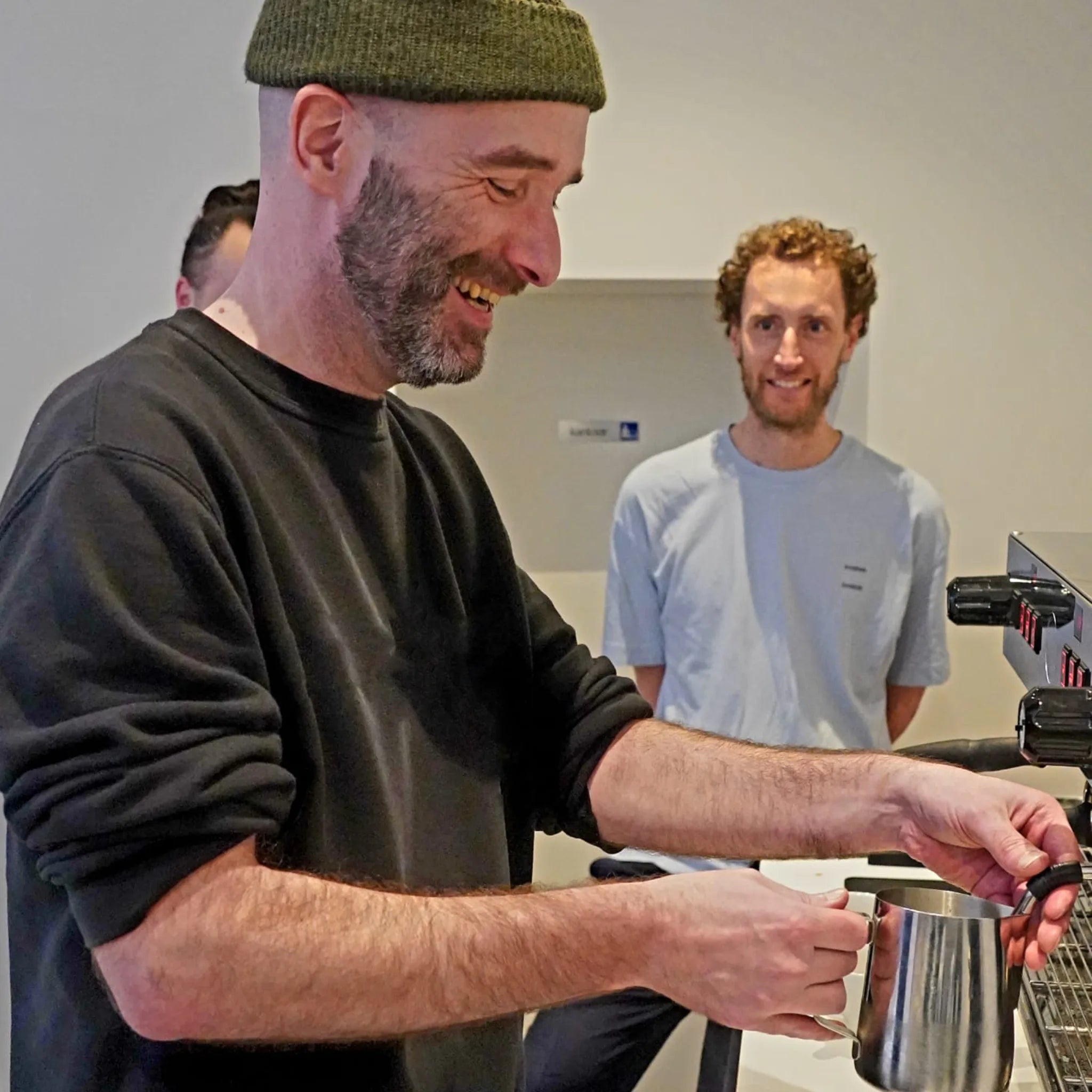 Coach en deelnemers lachen terwijl ze samen melk schuim technieken doornemen.