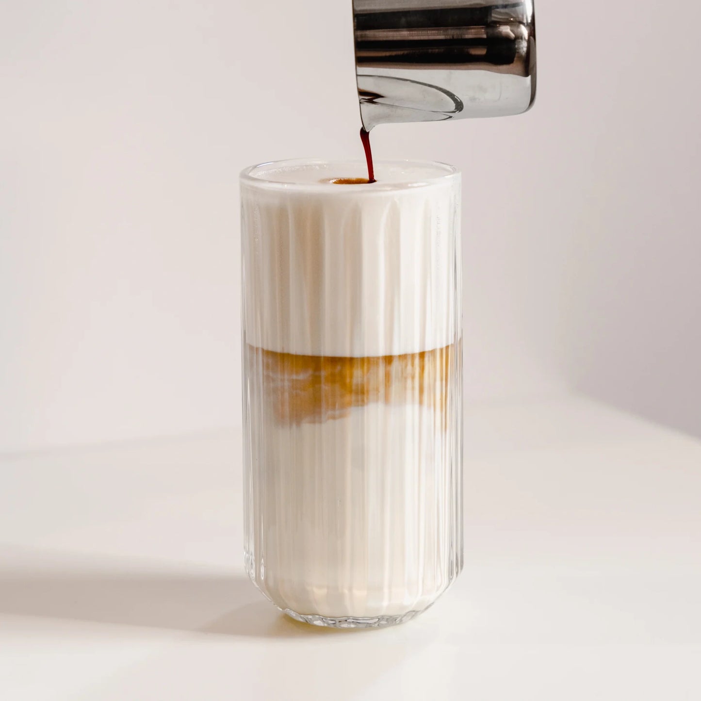 Latte macchiato in hoog geribbeld glas van 350 ml terwijl espresso door het melkschuim wordt geschonken