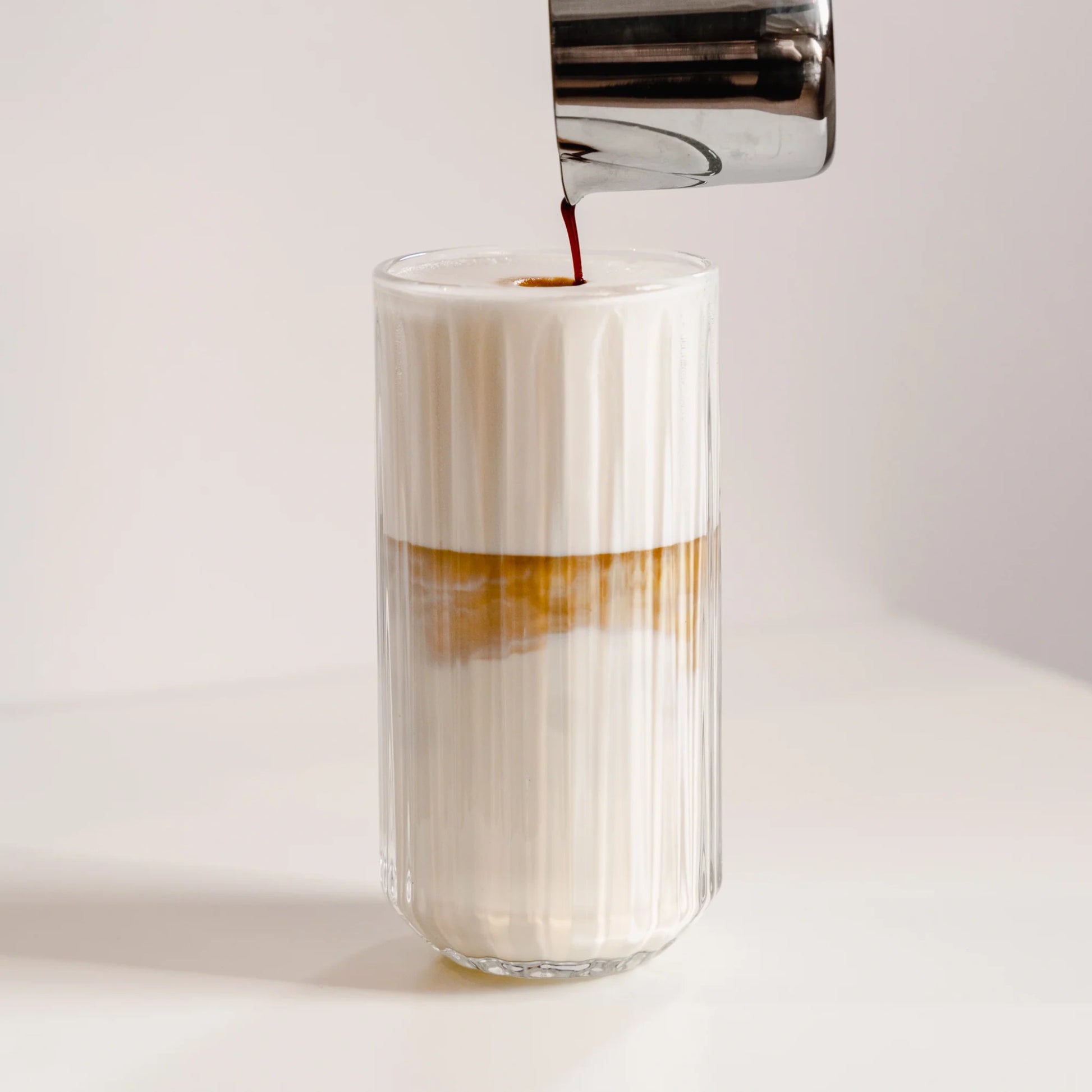 Latte macchiato in hoog geribbeld glas van 350 ml terwijl espresso door het melkschuim wordt geschonken