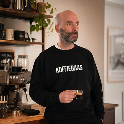 Man draagt zwarte KOFFIEBAAS sweater en houdt espresso vast in een warme thuisbarista-koffiehoek met espressomachine.