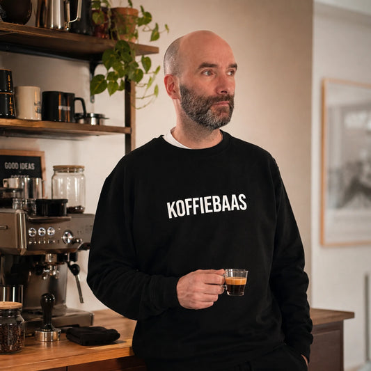 Man draagt zwarte KOFFIEBAAS sweater en houdt espresso vast in een warme thuisbarista-koffiehoek met espressomachine.
