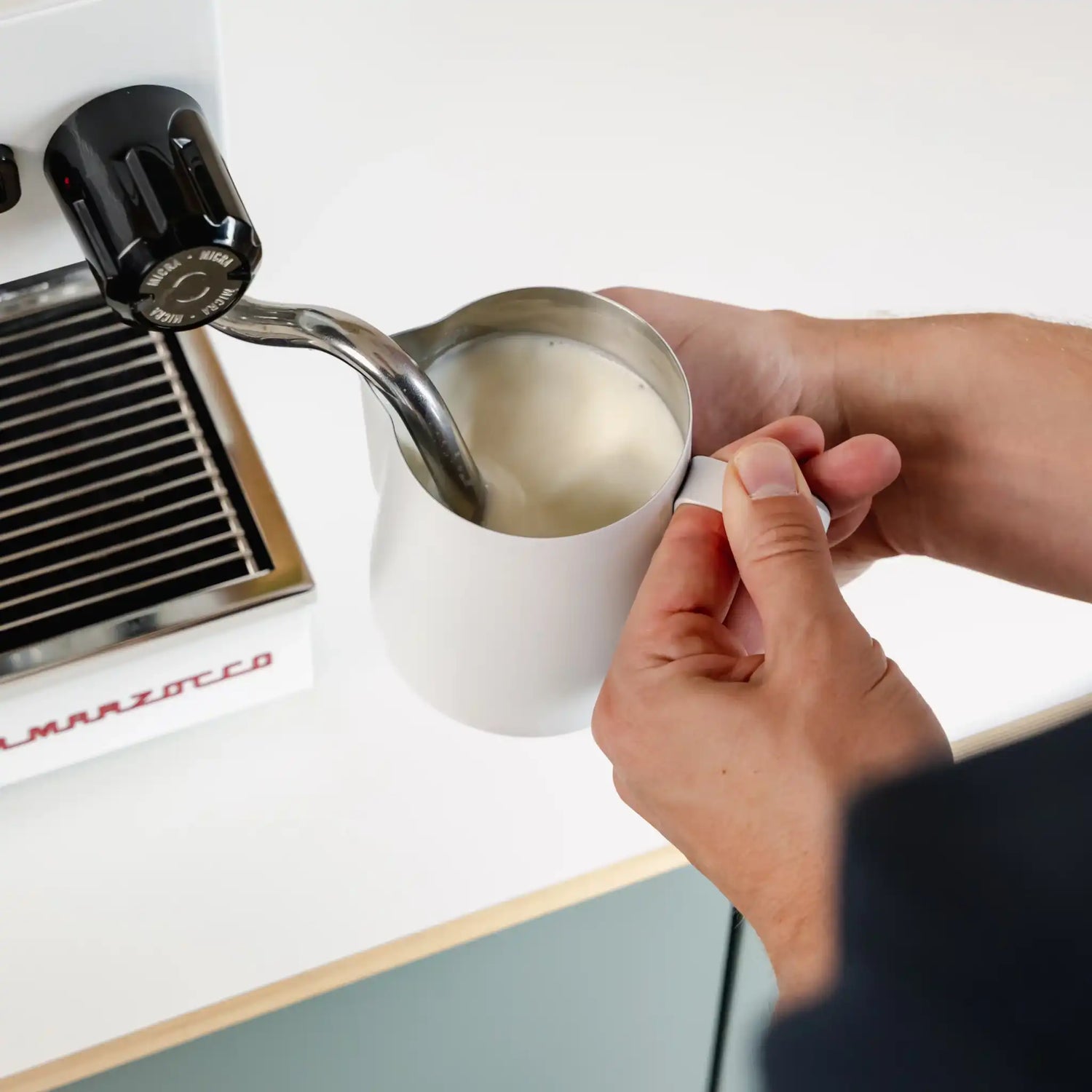 Thuisbarista schuimt melk op met witte melkkannetje onder stoompijp van espressomachine voor glad microschuim