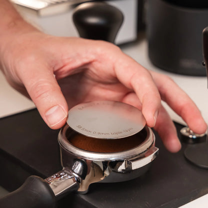 Hand plaatst een 58,5 mm puck screen op een aangetampte koffiepuck in een 58 mm E61-portafilter.