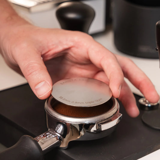 Hand plaatst een 58,5 mm puck screen op een aangetampte koffiepuck in een 58 mm E61-portafilter.