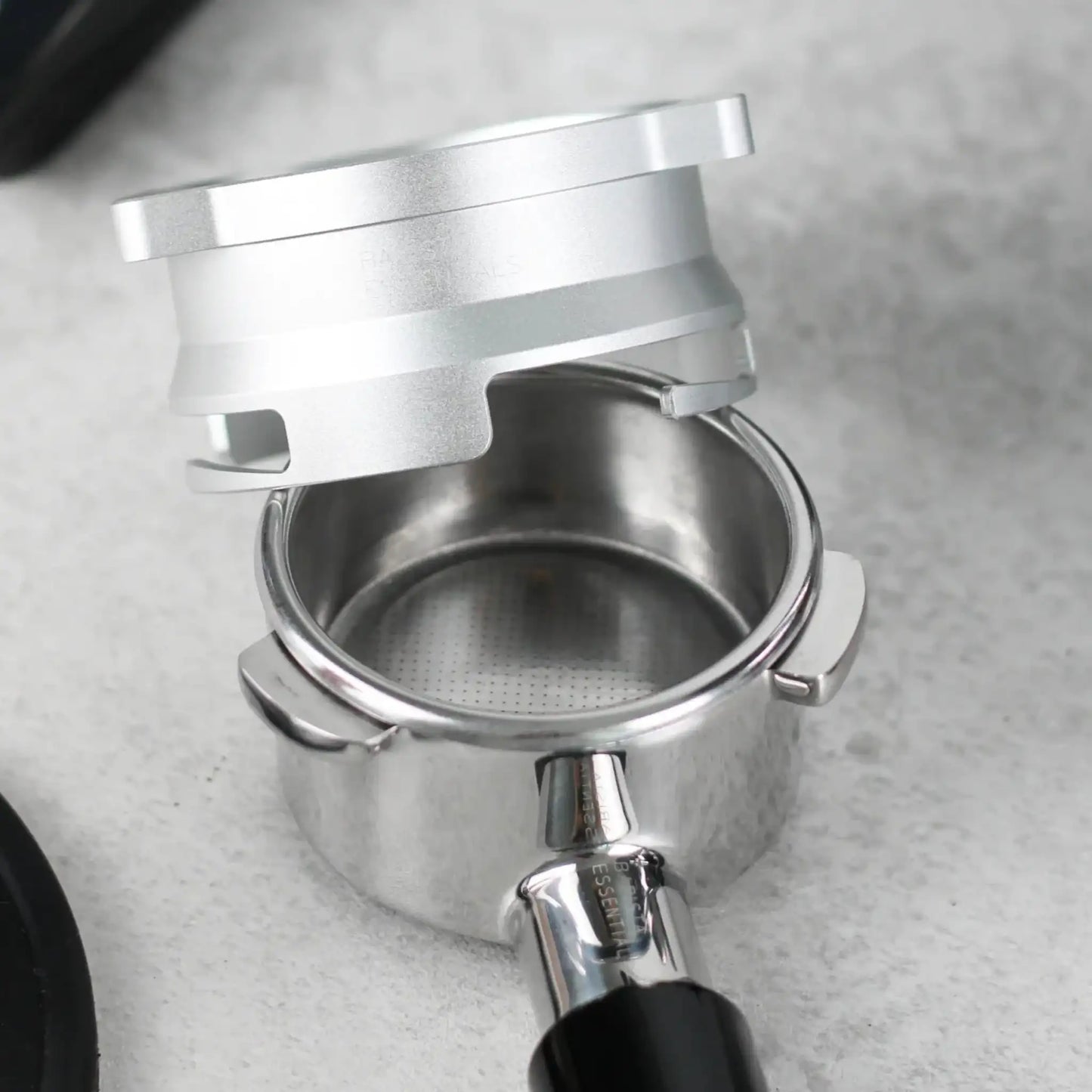 Nabije close-up van een lege portafilter op een lichtgrijze werkblad, met daarboven een zilverkleurige 53,3 mm doseerring klaar om koffie te vangen.