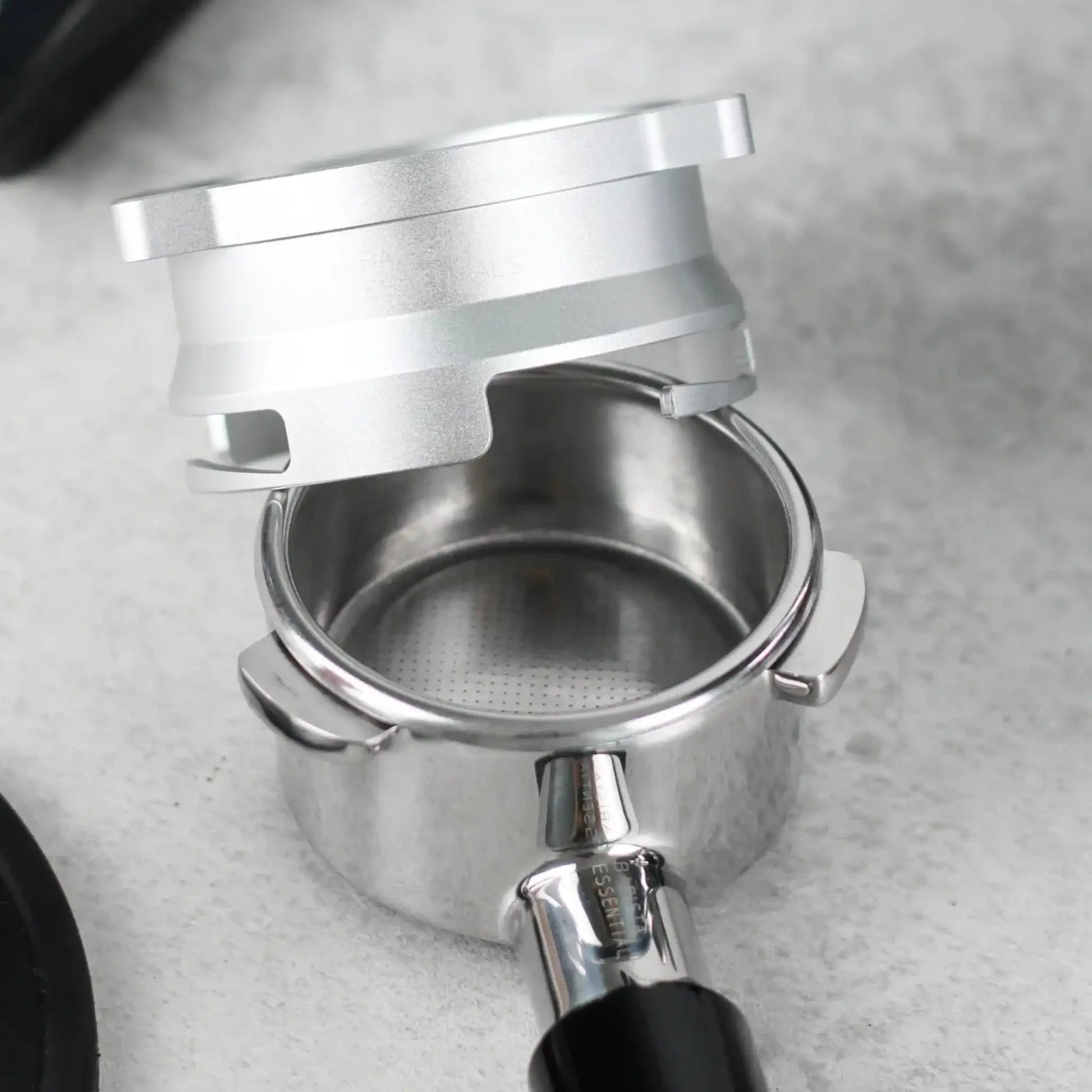 Nabije close-up van een lege portafilter op een lichtgrijze werkblad, met daarboven een zilverkleurige 53,3 mm doseerring klaar om koffie te vangen.