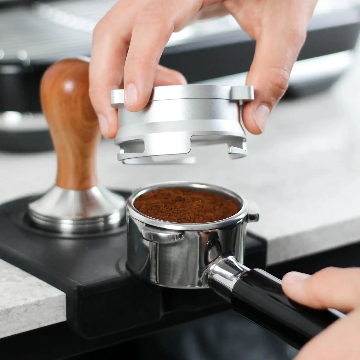 Hand plaatst een zilverkleurige 53,3 mm doseerring op een portafilter met versgemalen koffie, met een Sage Barista Express espressoapparaat en houten tamperset op de achtergrond.