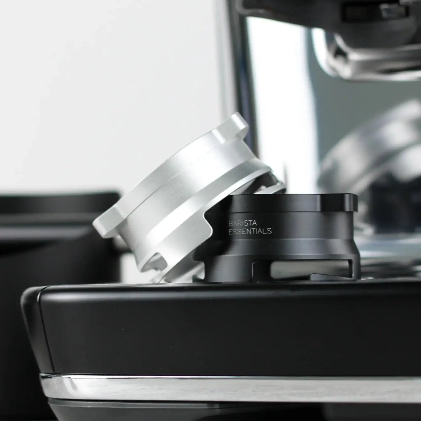 Een zilverkleurige en een zwarte 53,3 mm doseerring (Barista Essentials) liggen schuin tegen elkaar op de zijkant van een Sage Barista Express espressoapparaat.