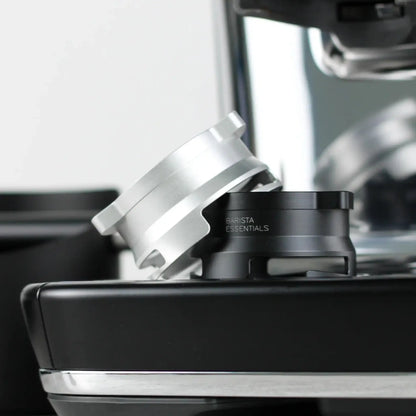 Een zilverkleurige en een zwarte 53,3 mm doseerring (Barista Essentials) liggen schuin tegen elkaar op de zijkant van een Sage Barista Express espressoapparaat.