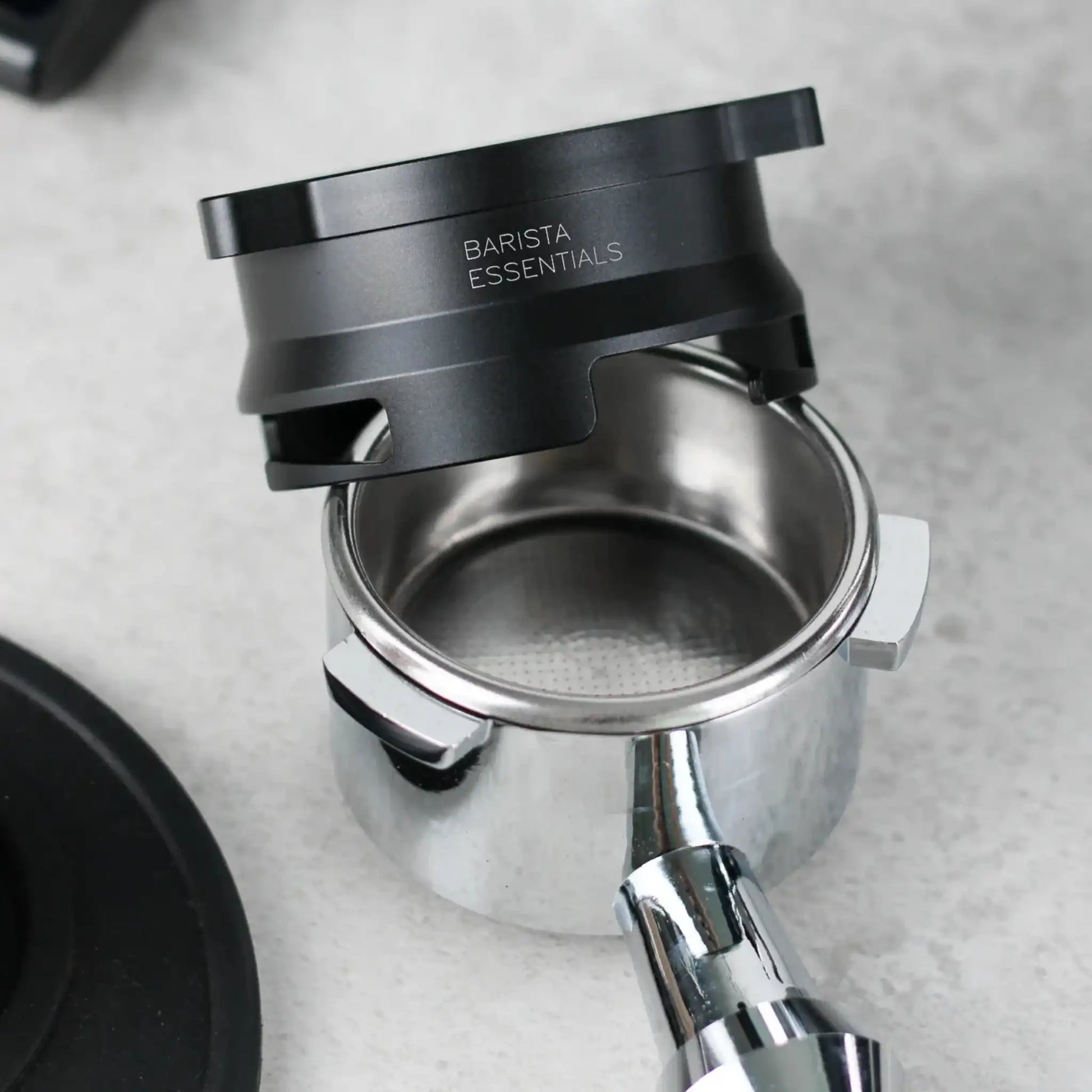 Zwarte 53,3 mm doseerring zweeft boven een lege portafilter op een grijze werkblad, klaar om gemalen koffie op te vangen.