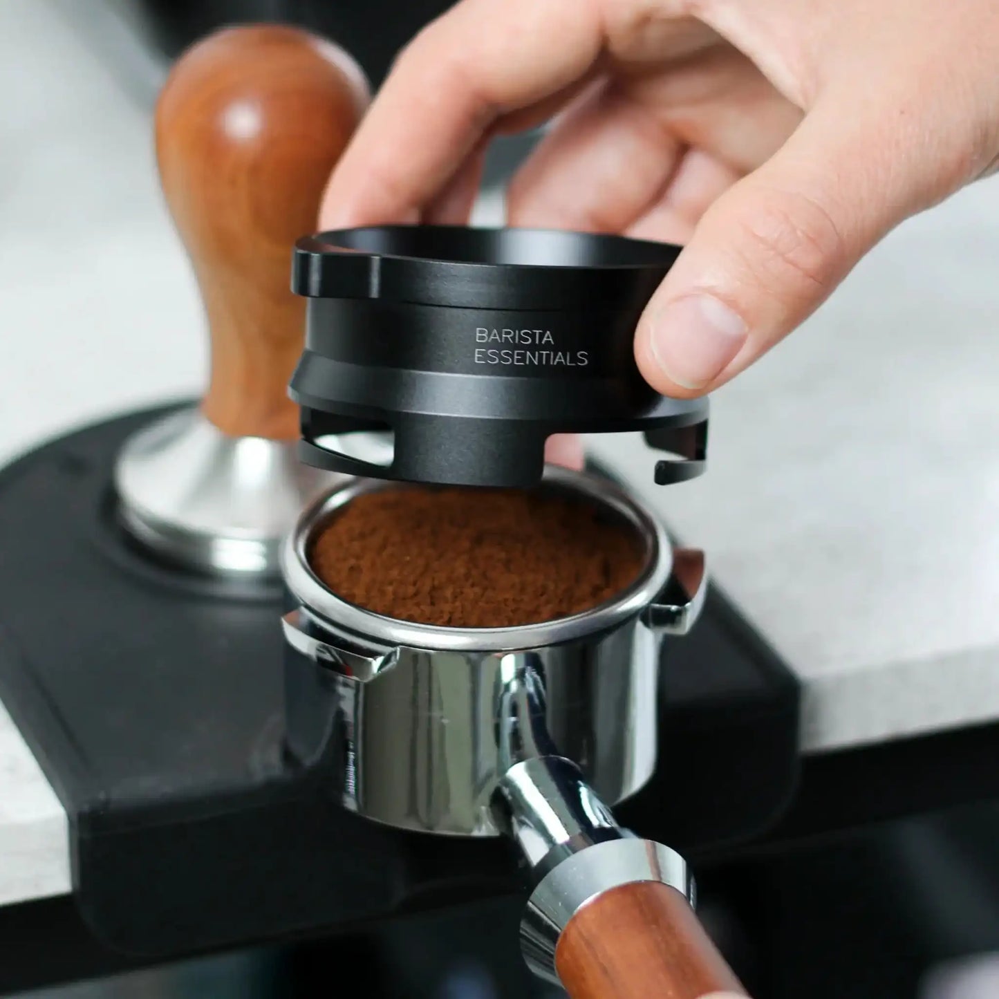 Hand plaatst een zwarte 53,3 mm doseerring op een portafilter met versgemalen koffie, met een Sage Barista Express en houten tamper op de achtergrond.