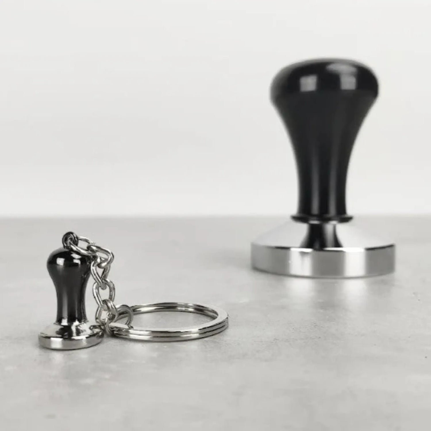 Mini tamper sleutelhanger van Barista Essentials naast een echte tamper – perfect koffiecadeau voor barista’s