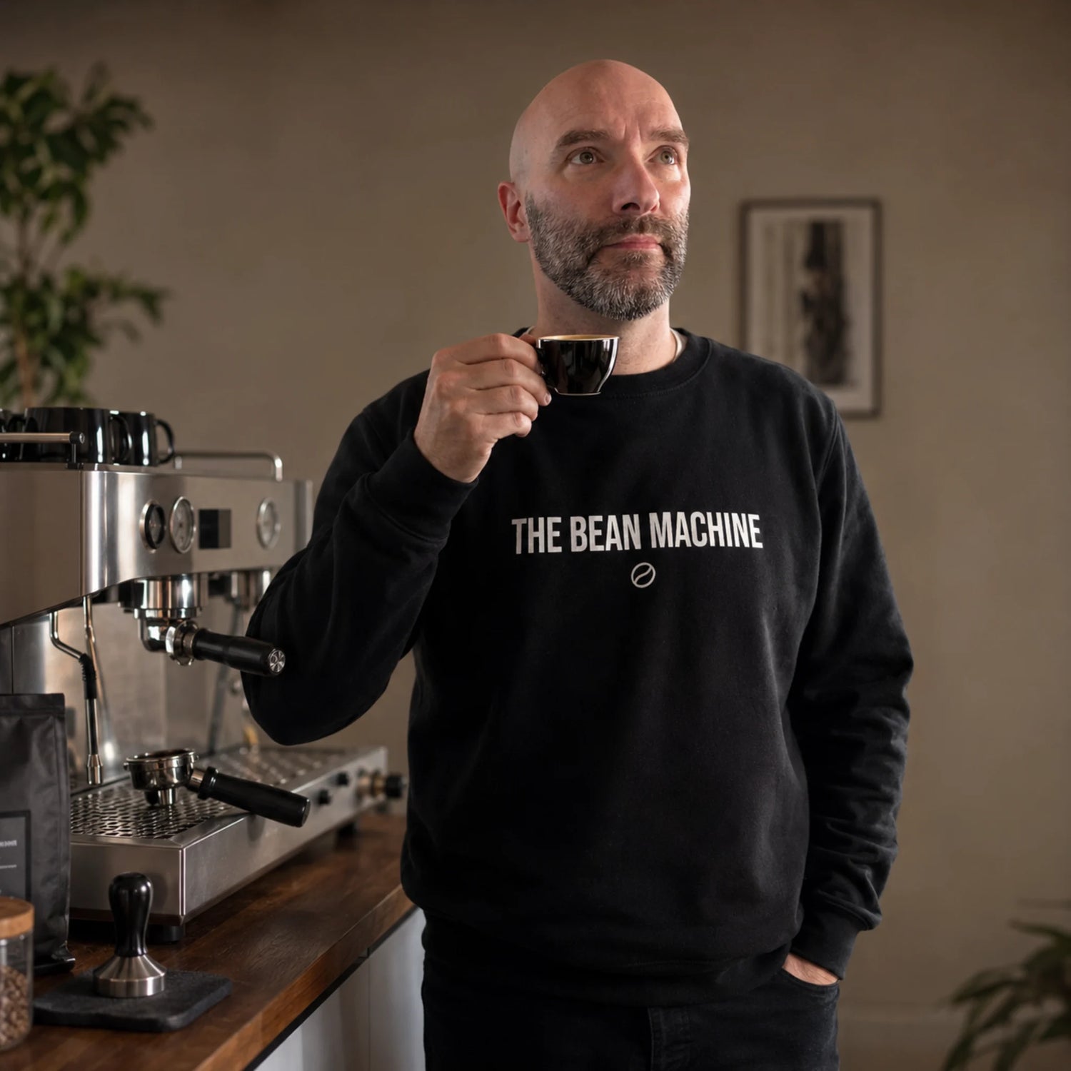 Barista draagt de zwarte The Bean Machine trui naast een espressomachine en geniet van een vers gezette espresso.