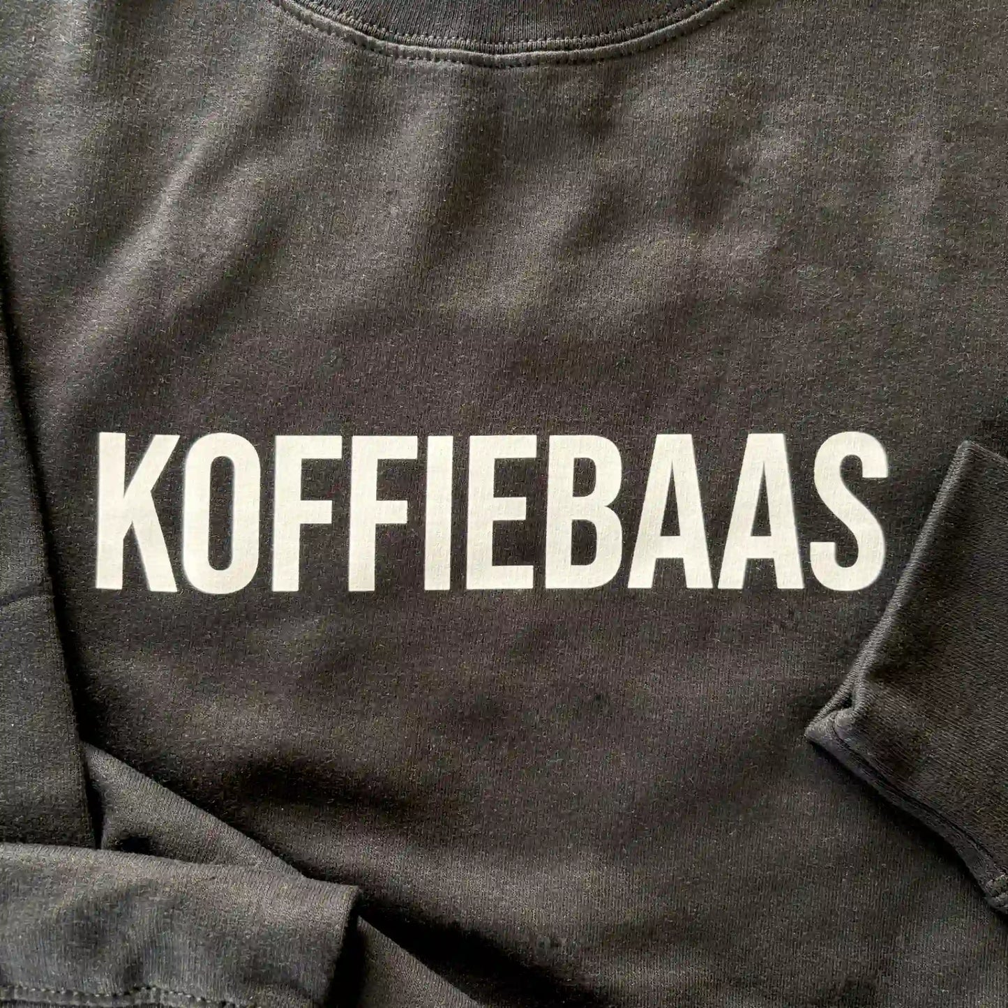 Close-up van de witte “KOFFIEBAAS” print op zwart barista sweatshirt