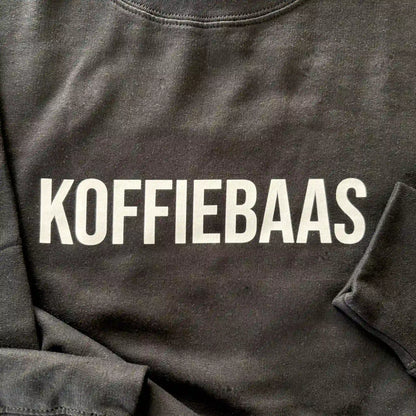 Close-up van de witte “KOFFIEBAAS” print op zwart barista sweatshirt