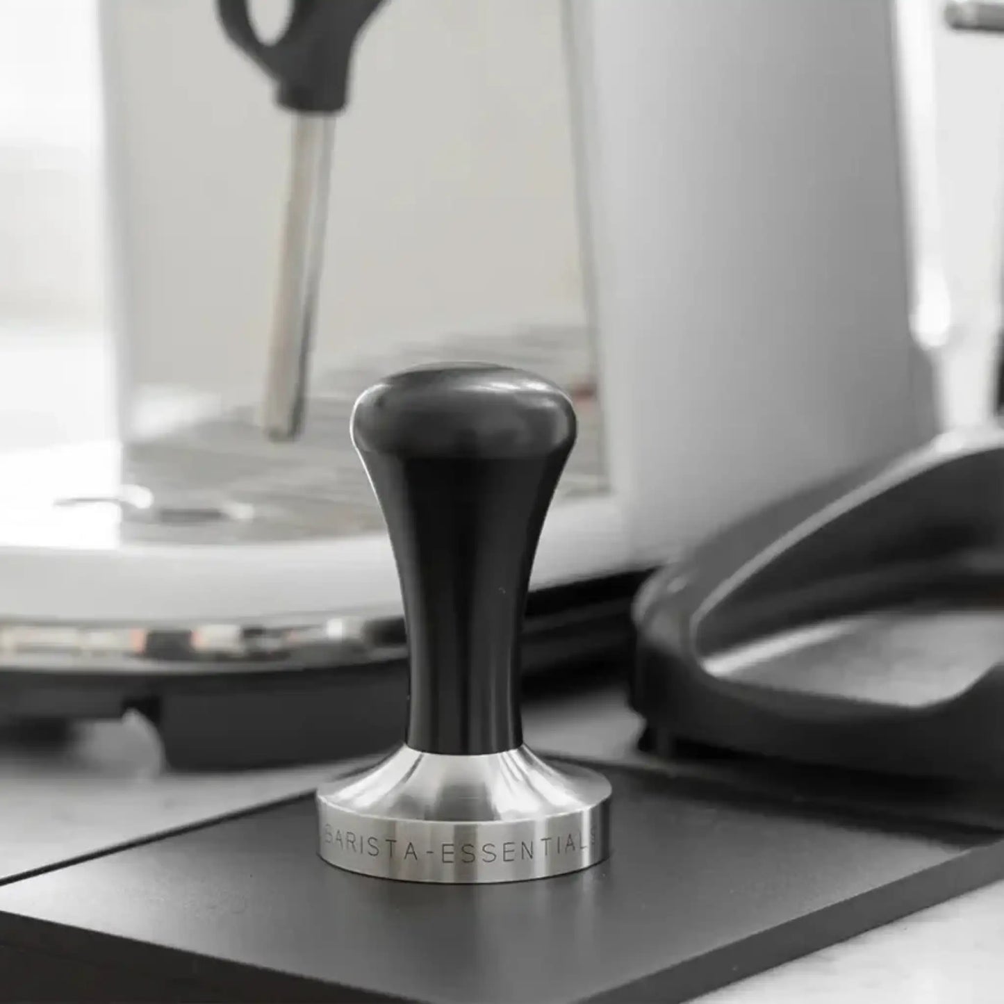 Zwarte espresso tamper met RVS basis op een tampermat naast een halfautomatische espressomachine.