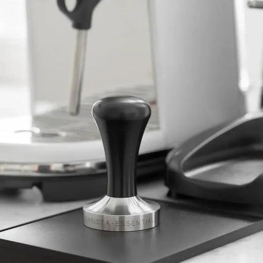 Zwarte espresso tamper met RVS basis op een tampermat naast een halfautomatische espressomachine.